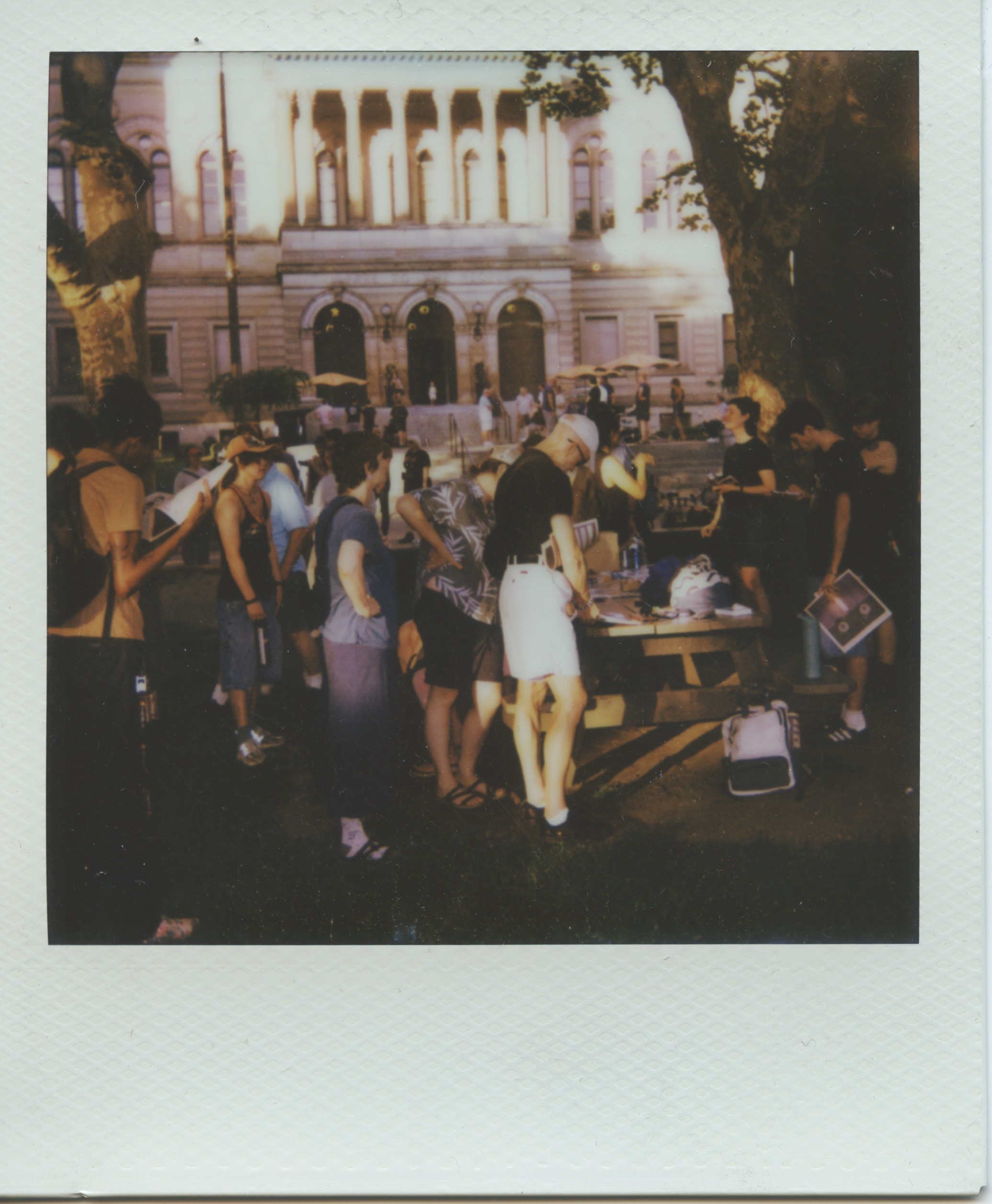 A large gathering under tree shade, people stand around a picnic table covered with photo albums and artists books filled with nightclub materials. In the background, three arched doorways to the Carnegie Library of Pittsburgh Oakland Main Branch are brightly lit by the sun.